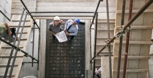 Male architect and male project manager discussing construction progress standing at bottom of scaffold staircase at construction site.